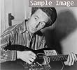 1943 Woody Guthrie, half-length portrait, seated, facing front, playing a guitar that has a sticker