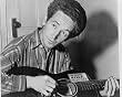 1943 photo Woody Guthrie, half-length portrait, seated, facing front, playing a guitar that has a st